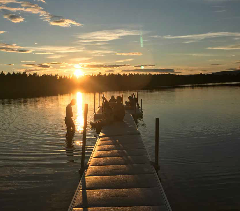 Bild av en brygga som går ut i en stilla sjö. Solen är på väg ner. Folk badar på bryggan. 