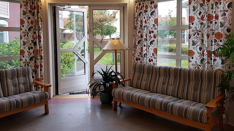 En soffa framför ett fönster med färgglada gardiner. En balkongdörr står öppen  och leder ut mot en innergård.