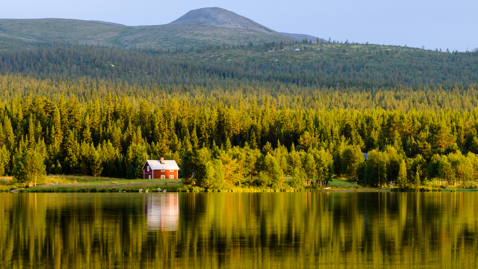 Ett rött hus vid en sjö, i bakgrunden skog och ett fjäll. Sommartid.