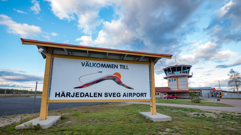Skylt på gräsmatta med texten Välkommen till Härjedalen Sveg airport framför ett staket och en byggnad med ett trafiktorn.