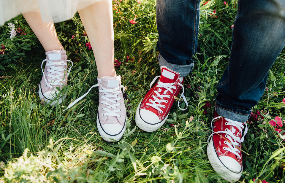 Två par fötter i rosa och röda lätta sommarskor med knytning står i grönt gräs. En person är barbent och har en flortunn kjol, en person har blå jeans som är uppvikta nertill.