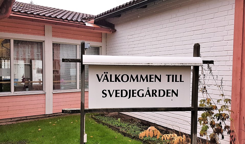 En vit tegelvägg och i vinkel en rosa vägg med stora fönster. Framför huset på gräsmattan en skylt med texten Välkommen till Svedjegården.