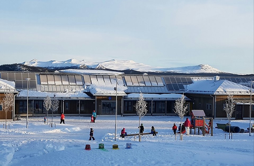 Barn leker på en snöig skolgård framför en förskola. I bakgrunden snöklädda fjäll.
